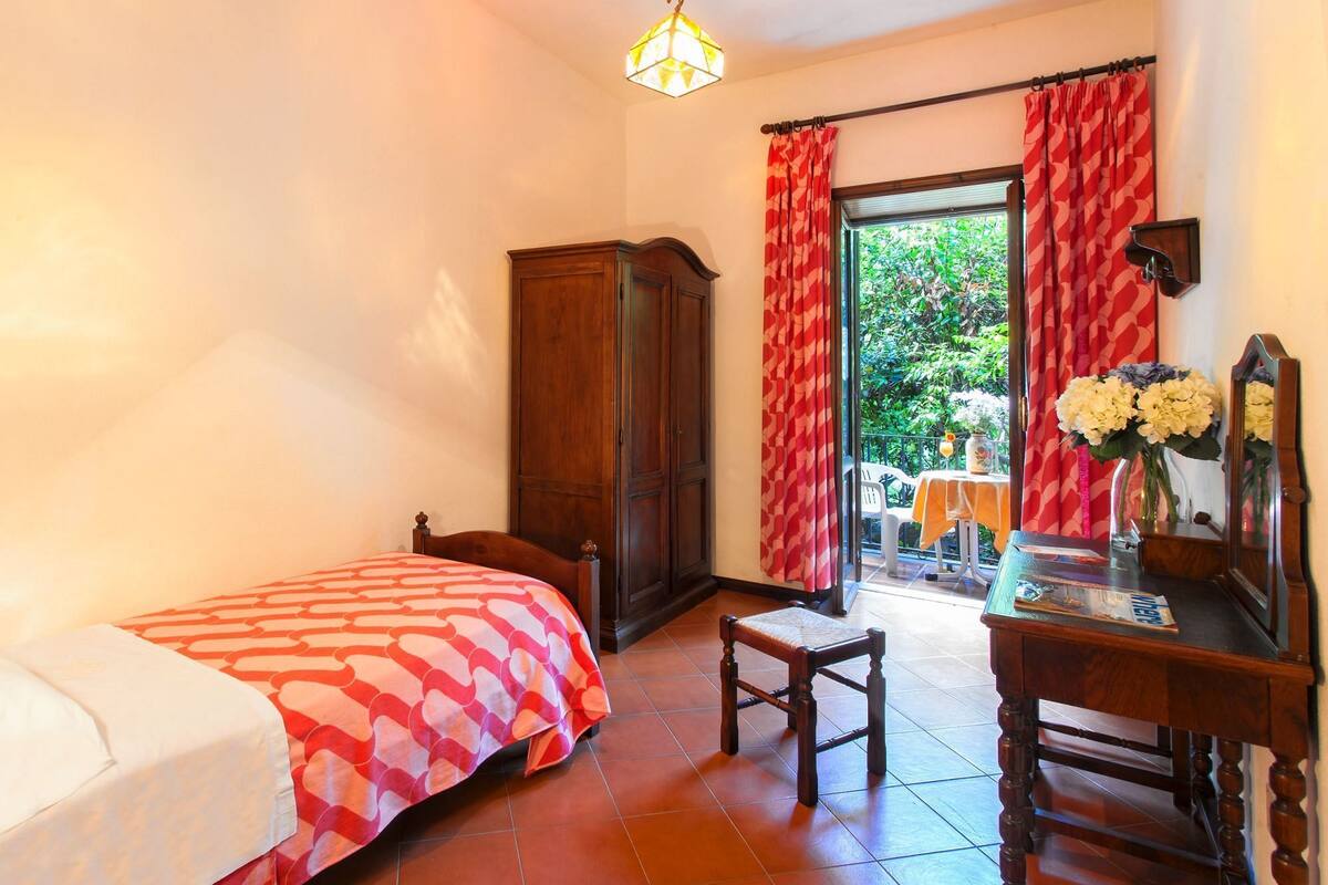 A bedroom with a bed, a wooden wardrobe, a desk with a chair, and a window with red curtains.