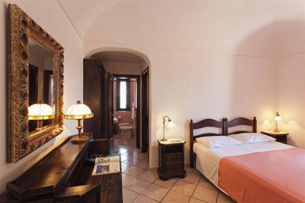 A hotel room with a bed, a desk with a lamp, a mirror, and a small nightstand with a lamp.