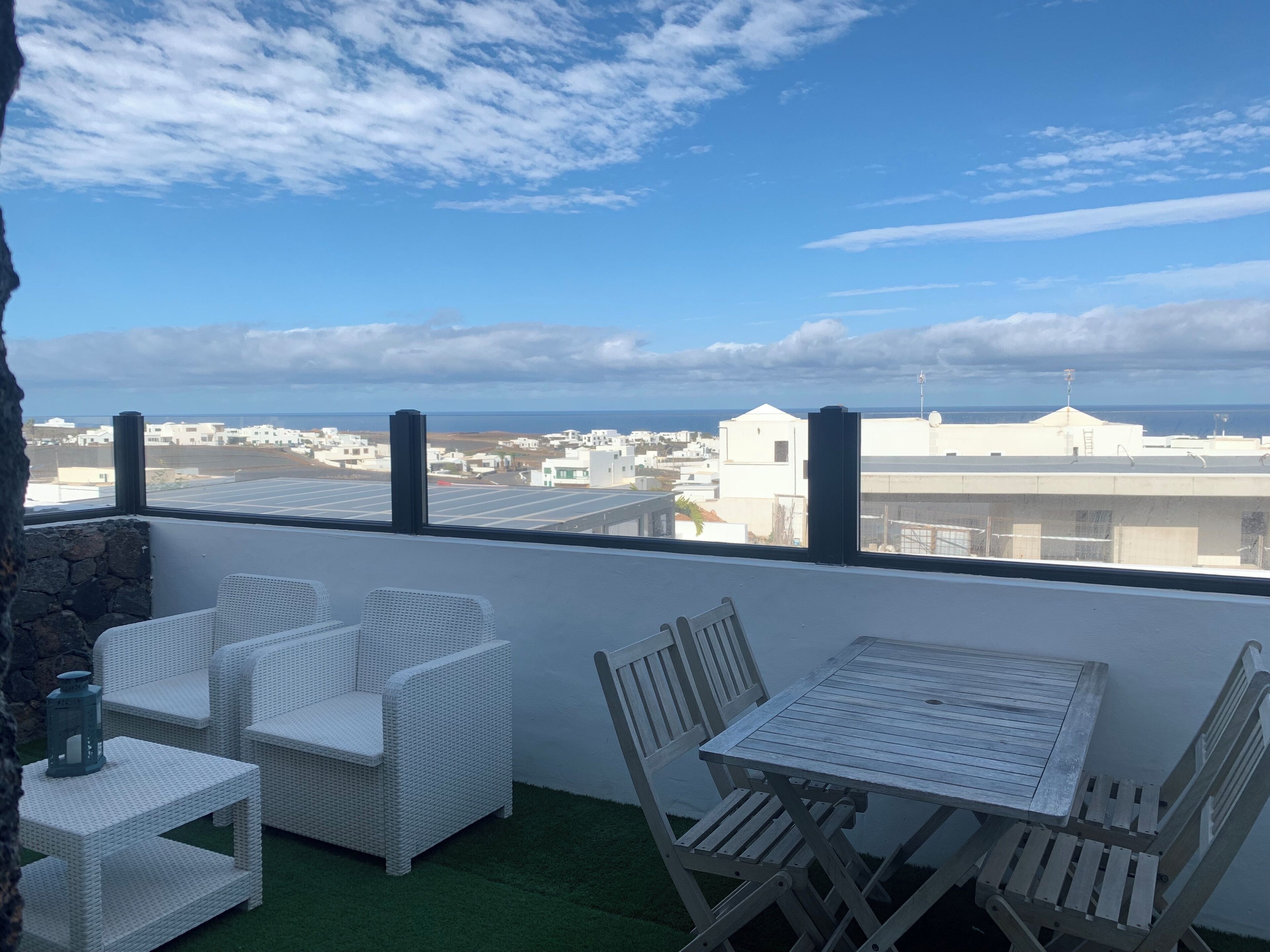 Un balcon doté de meubles en osier blanc, d’une table et de chaises, offrant une vue sur une ville côtière avec ses bâtiments et la mer.