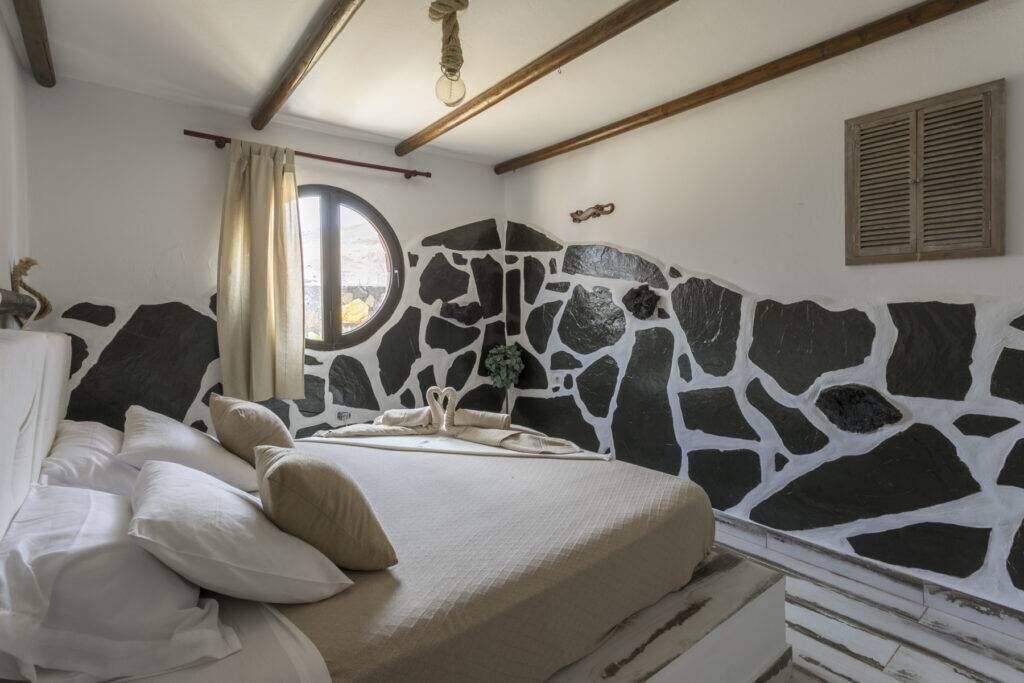 Ein Schlafzimmer mit Steinwand, einem Bett mit beige farbenem Bettzeug, einem Fenster mit Vorhängen und einem Holzbalken an der Decke.