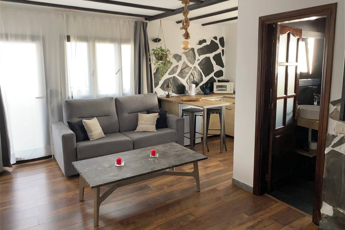 Un salon avec un canapé gris, une table basse en bois et un coin cuisine doté d’un pare-feu en pierre.