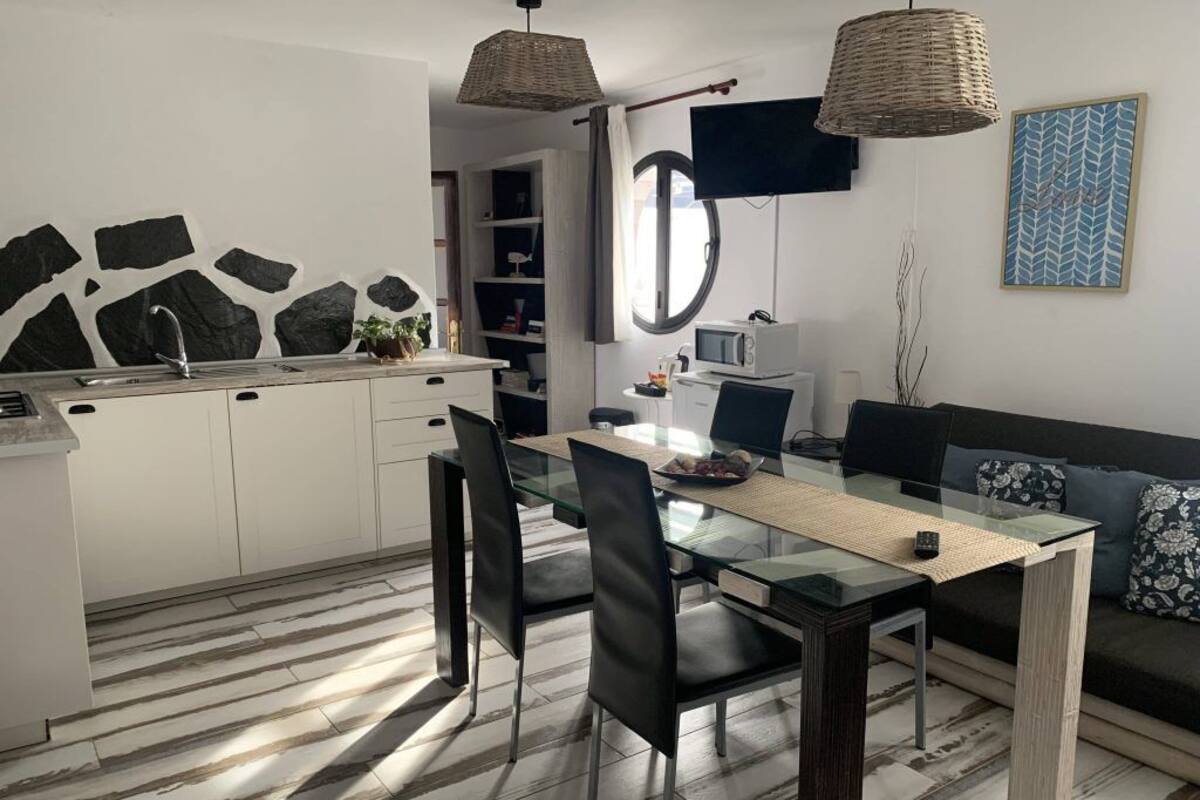 Une cuisine moderne avec des meubles blancs, une table à manger avec des chaises, un tél éviseur fixé au mur et un canapé avec des coussins.