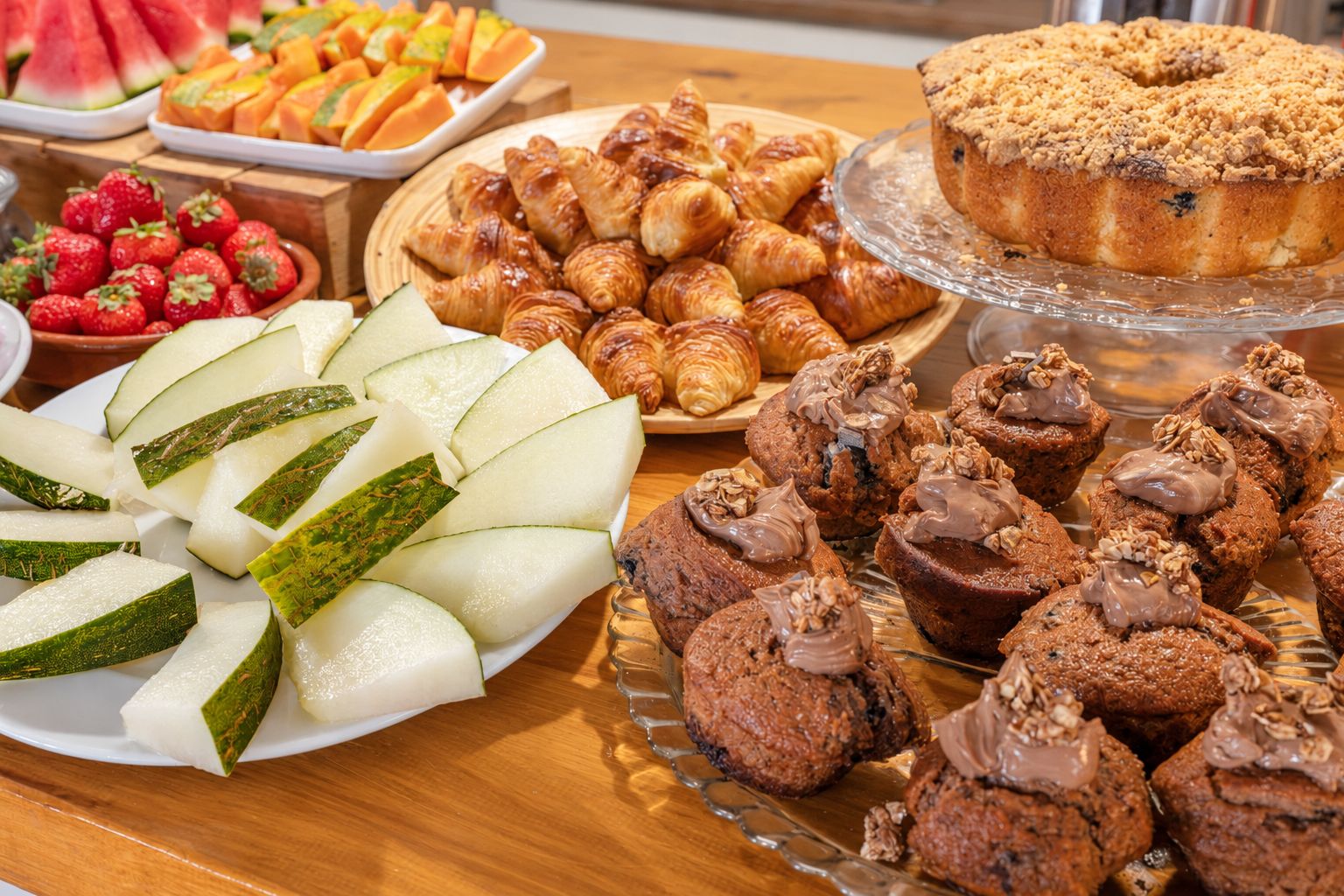 A variety of food items including sliced cucumbers, strawberries, croissants, muffins, and a bundt cake on a wooden table.