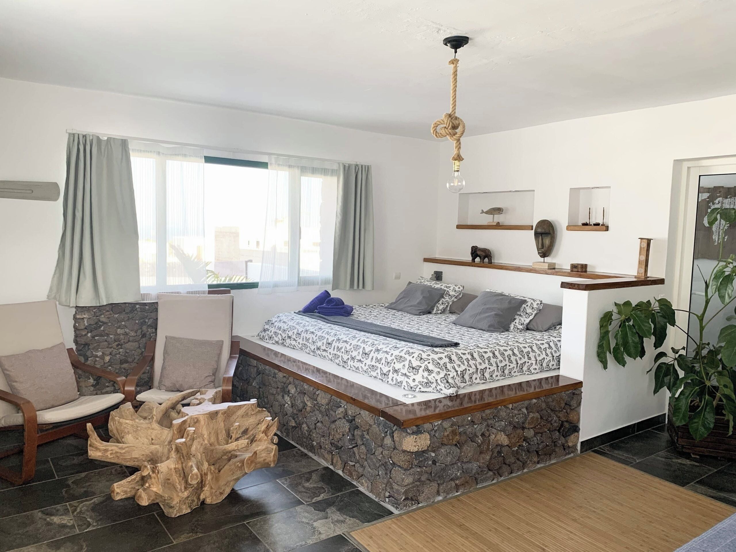 A modern bedroom with a stone bench, a bed with patterned bedding, a wooden headboard, and a large window with curtains.