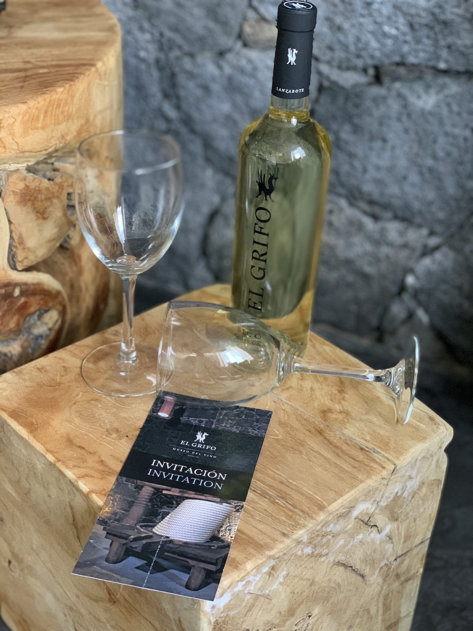 Una botella de vino El Grifo, dos copas de vino vacías y una tarjeta de invitación sobre un bloque de madera.