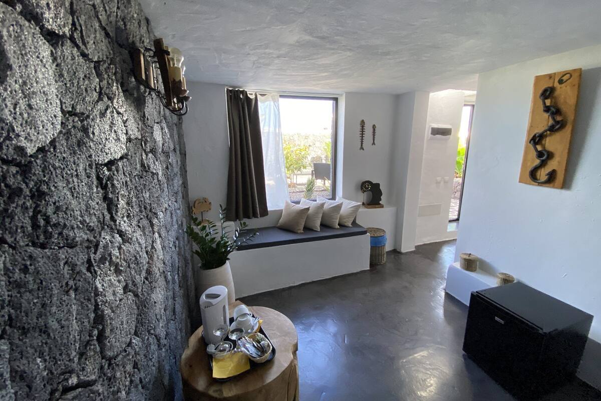 Une pièce avec un mur en pierre, un banc en bois avec des coussins, une fenêtre avec des rideaux, une sculpture fixée au mur et un meuble noir.