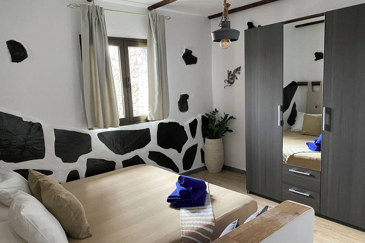 Une chambre à coucher avec un lit, un miroir, une plante en pot et une fenêtre avec des rideaux.