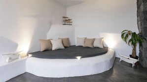 Una cama redonda con colchón gris y almohadas color beige, rodeada de paredes blancas y una pequeña planta en maceta.