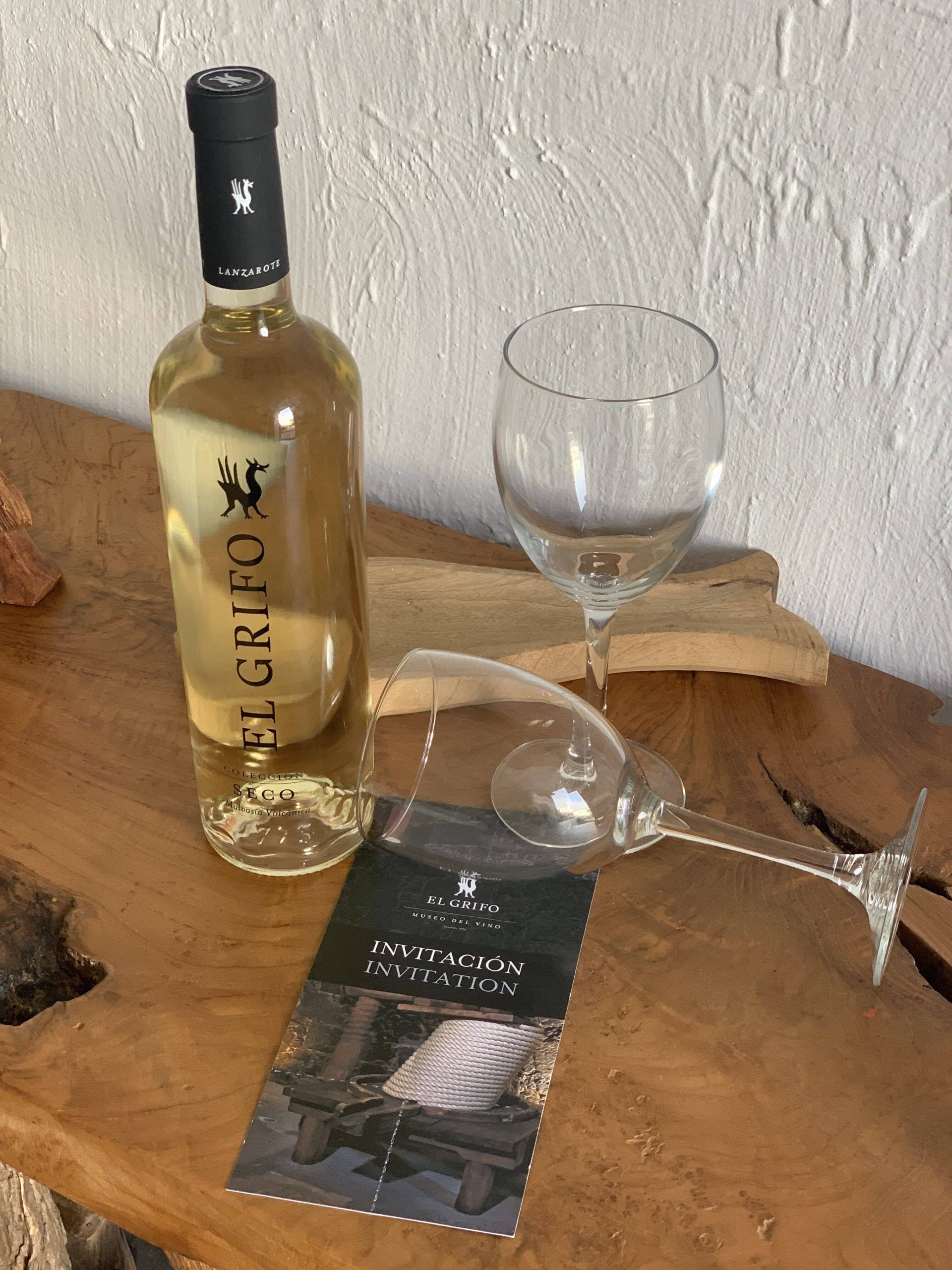 A bottle of El Grifo wine, a glass, and an invitation card on a wooden surface.
