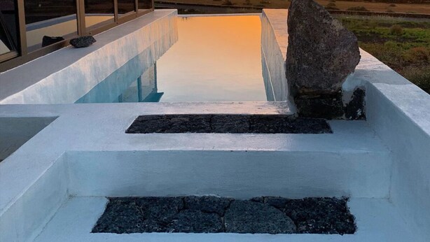 Piscina moderna al aire libre con una roca como pieza central, rodeada por una estructura blanca con ventanales grandes, frente a una puesta de sol y montañas distantes.