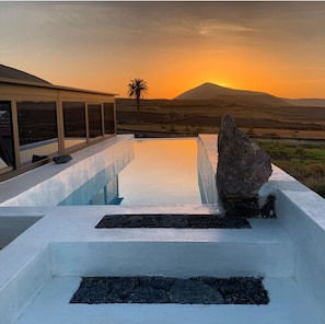 A modern outdoor pool with a rock centerpiece, surrounded by a white structure with large windows, set against a sunset and distant mountains. - Finca Marisa (Tinajo)