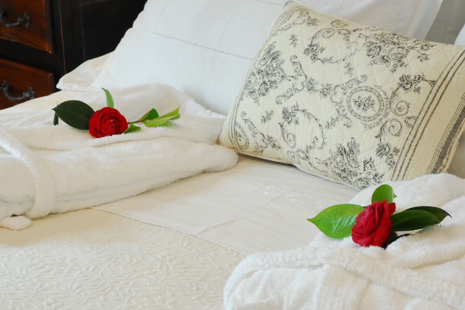 Una cama bien hecha con sábanas blancas y dos rosas rojas sobre los almohadones.