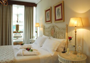 A hotel room with a neatly made bed, a round bedside table with a lamp, a small table with a vase of flowers, and a window with curtains.