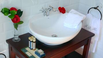 Ein Badezimmer mit einem weißen Waschbecken, einem Spiegel und einer Holzkommode. Auf der Kommode stehen rote Rosen in einer Vase, ein Handtuch mit Initialen und eine Lotionflasche.
