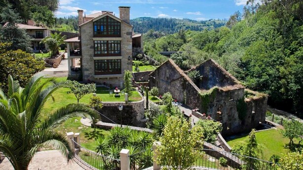 A large stone building with a balcony, surrounded by lush greenery and a mountainous backdrop. - Rusticae Torre do Río (Caldas de Reis)