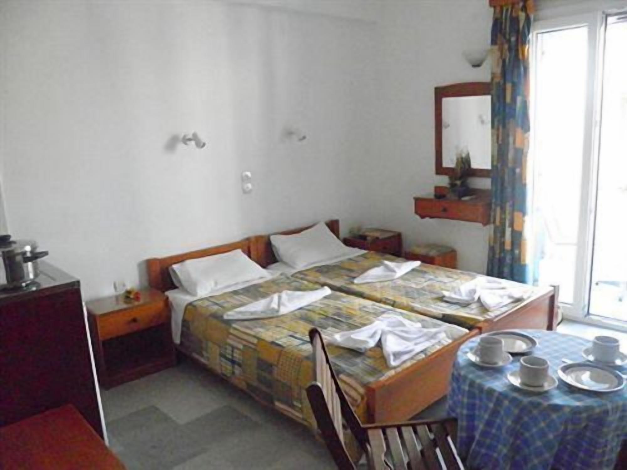 A hotel room with two beds, a small table with plates and cups, a desk with a kettle, and a window with curtains.