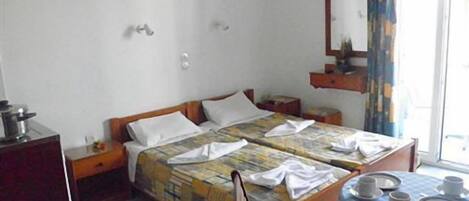 A hotel room with two beds, a small table with plates and cups, a desk with a kettle, and a window with curtains.