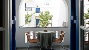 A room with a round table and two wicker chairs, a window with an arched frame, and a view of buildings outside. - Rea Sun Hotel Studios Apartments (Naxos)