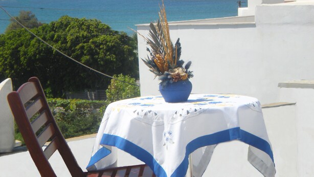 A table with a white tablecloth and blue trim, a potted plant, and a wooden chair. A coastal view with mountains and the sea is visible in the background. - Rea Sun Hotel Studios Apartments (Naxos)
