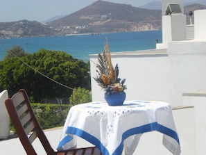 Ein Tisch mit weißem Tuch und blauer Bordüre, ein Blumentopf und ein Holzstuhl. Im Hintergrund ist eine Küstenlandschaft mit Bergen und dem Meer zu sehen.