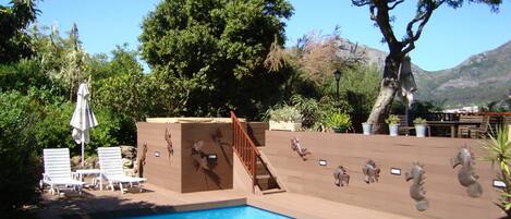 A pool area with a wooden deck, lounge chairs, and a staircase leading to a covered area with plants.