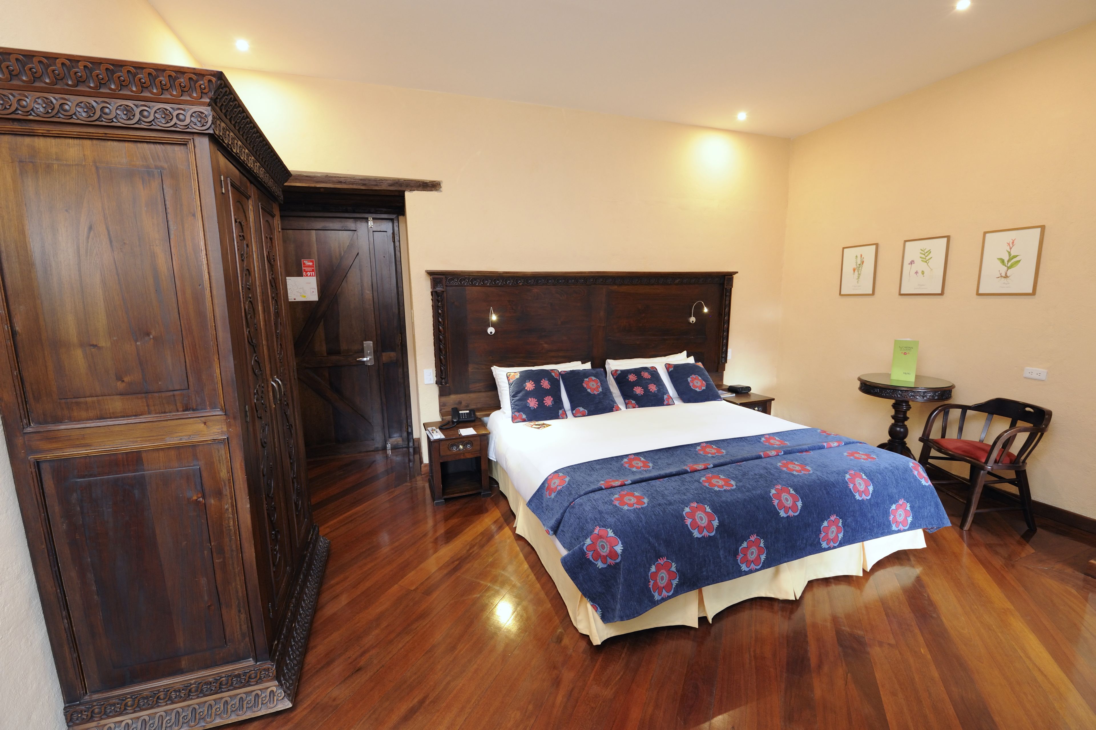 A hotel room with a large wooden wardrobe, a bed with a floral bedspread, a nightstand, a small table with a chair, and framed botanical prints on the wall.