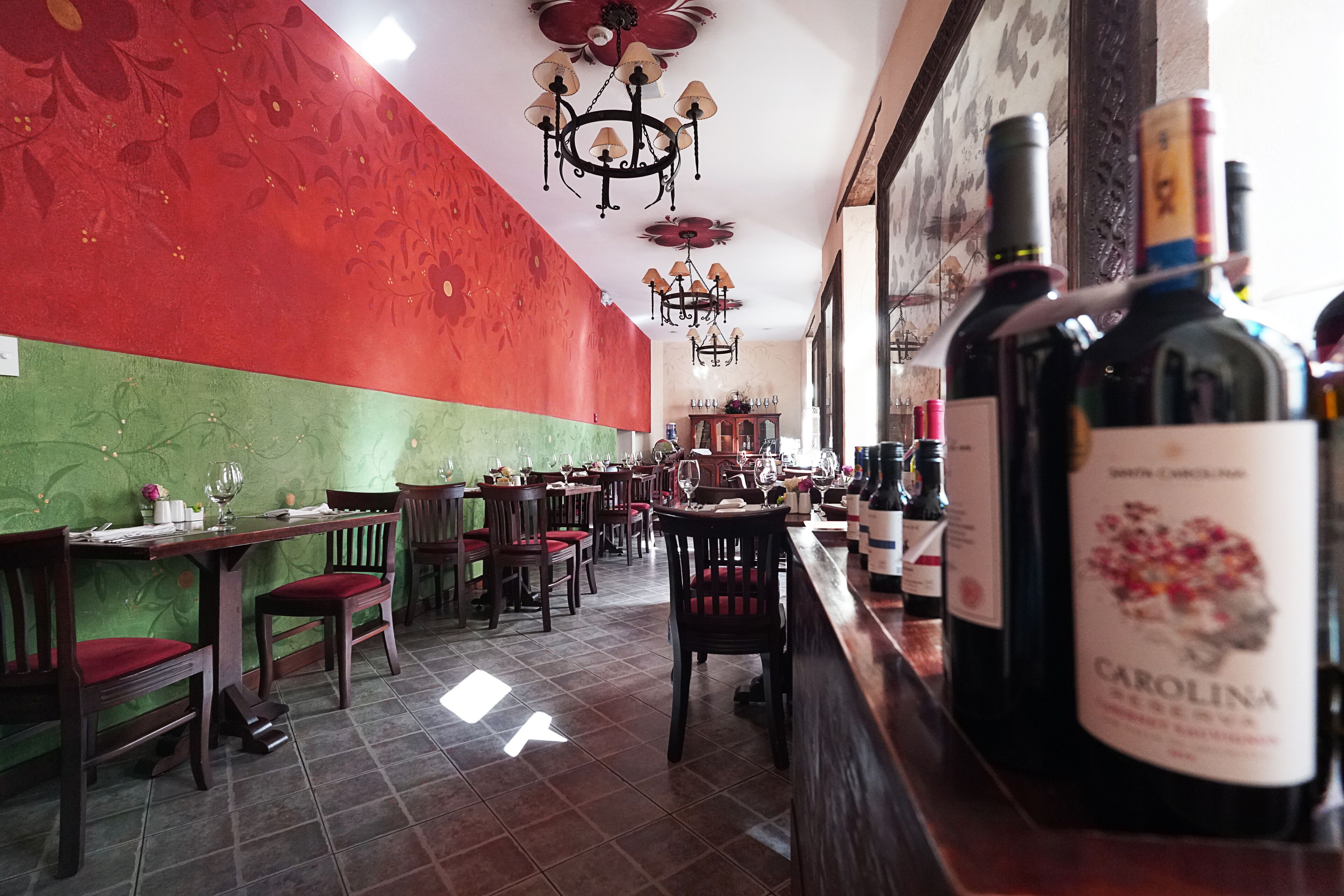 A restaurant interior with red and green walls, wooden tables and chairs, and a bar area with wine bottles.