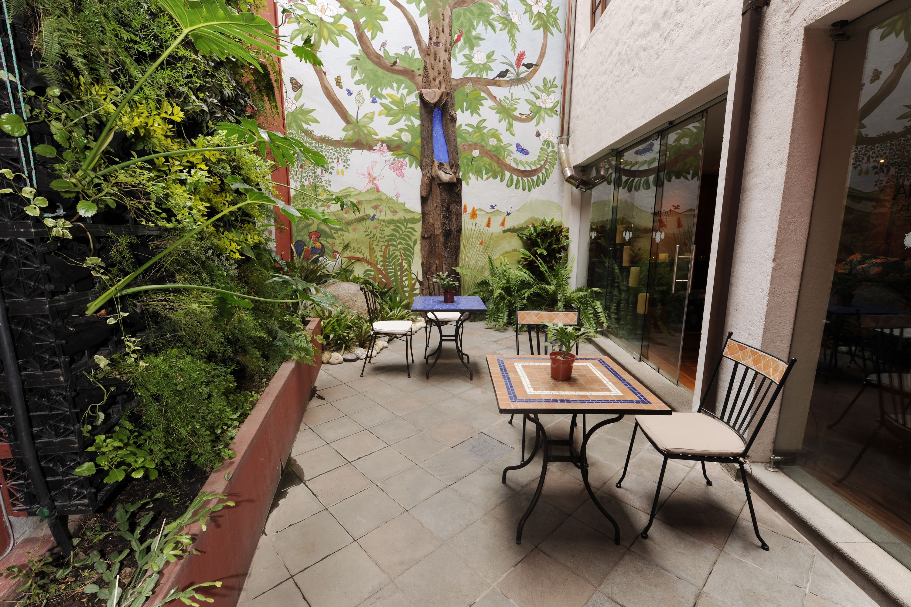 Um pátio externo com mesa e cadeiras, cercado por vegetação exuberante e um mural de uma árvore.