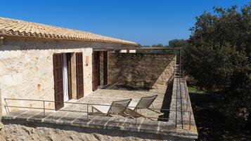 A stone building with a tiled roof, a covered outdoor seating area with chairs, and a stone wall.