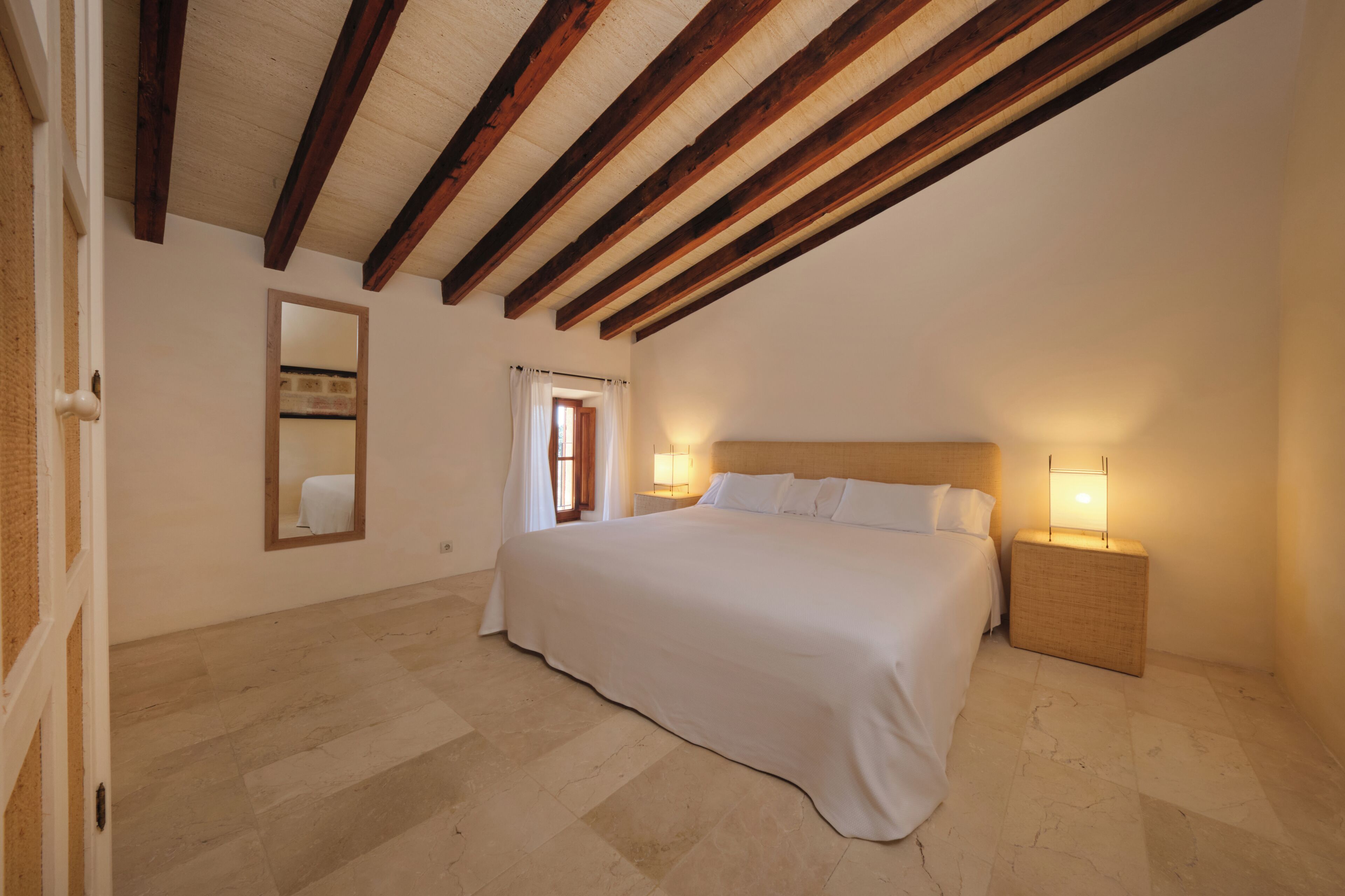 A bedroom with a large bed, wooden beams on the ceiling, a nightstand with a lamp, and a mirror on the wall.