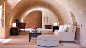 A cozy room with a stone vaulted ceiling, a white sofa, a wooden bench, a small table with a vase of flowers, and a lamp.