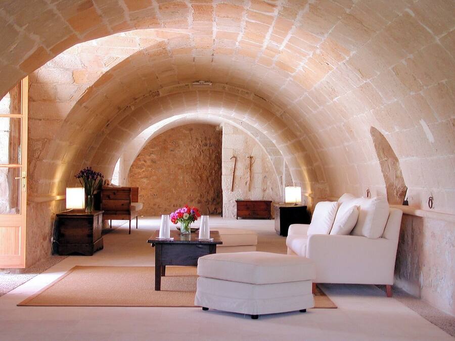 A cozy room with a stone vaulted ceiling, a white sofa, a wooden bench, a small table with a vase of flowers, and a lamp.