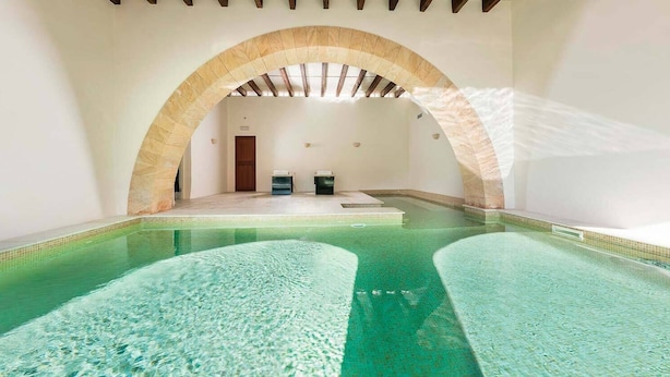 An indoor swimming pool with clear water, a stone archway, and a small seating area with two chairs.