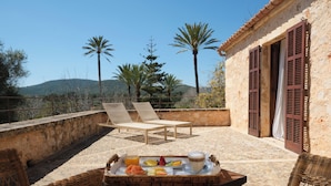 A terrace with wicker furniture, a tray of breakfast, and a view of palm trees and hills.