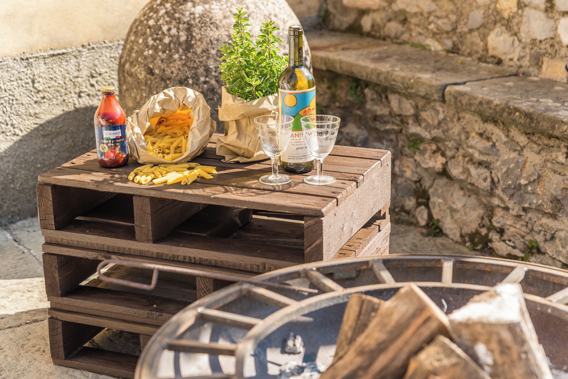 A rustic outdoor setting with a wooden table holding pasta, wine bottles, and glasses.