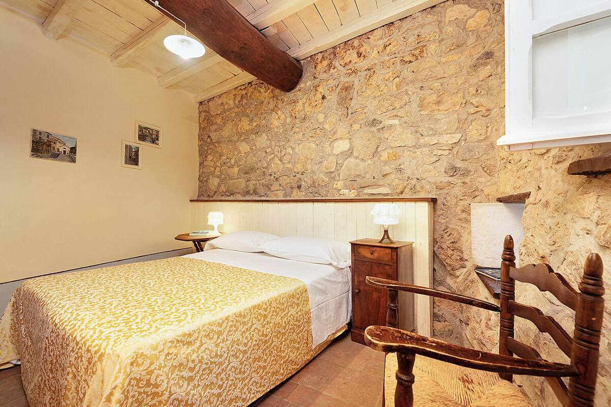 Un dormitorio con pared de piedra, una cama con colcha estampada, dos lámparas de noche, una silla de madera y una mesita.