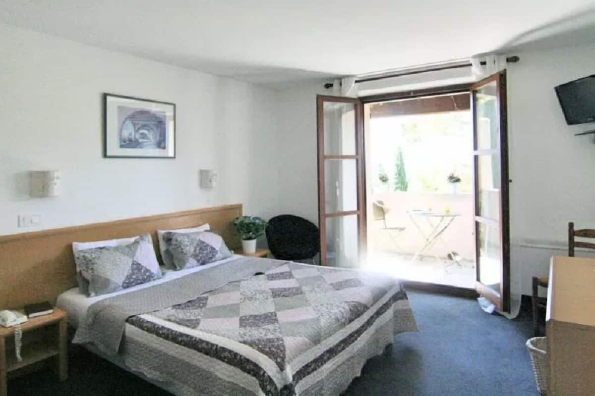 A bedroom with a bed, a bedside table, a chair, a small table with a plant, and a sliding glass door leading to an outdoor area.