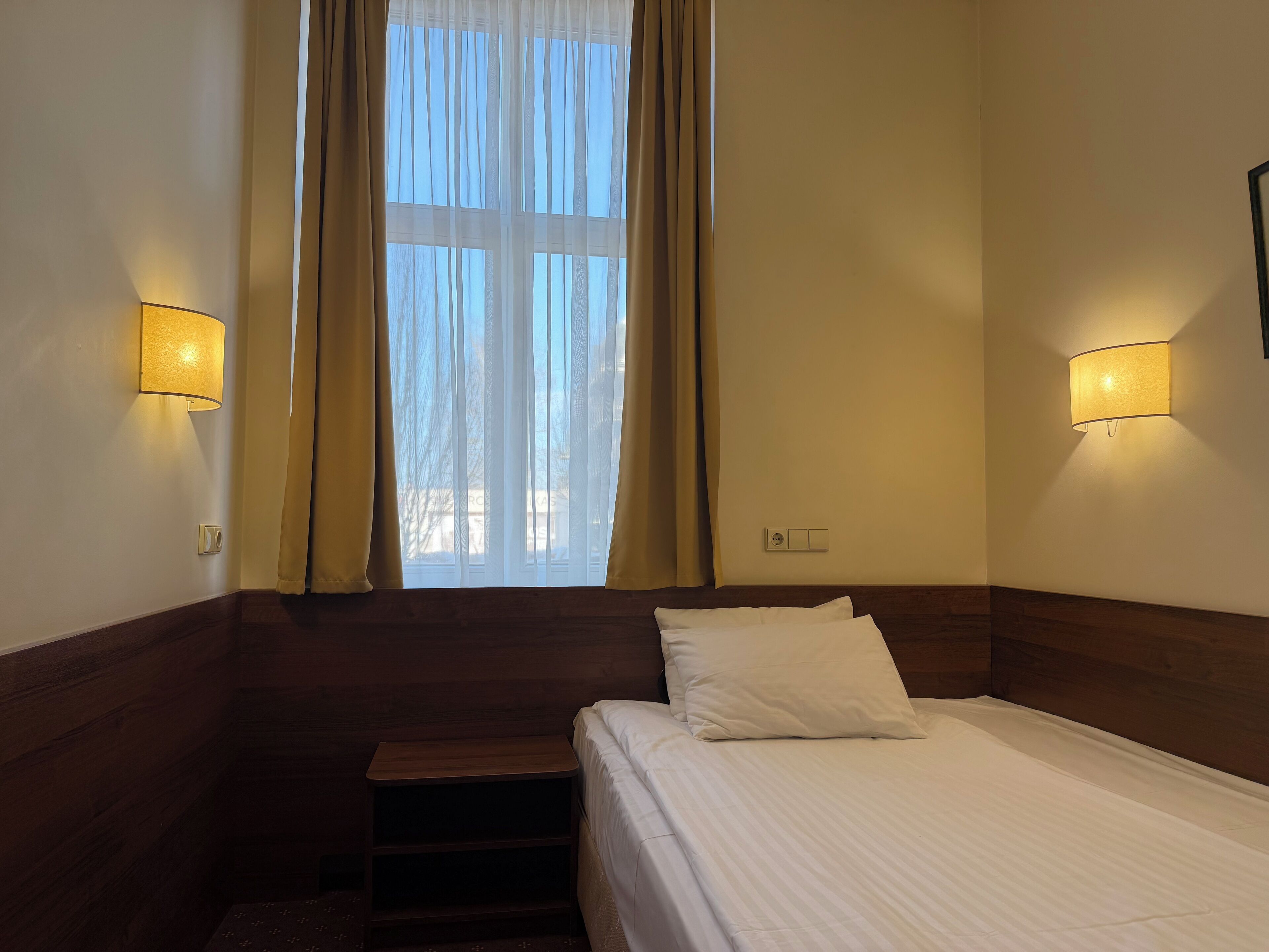 Una habitación de hotel con cama, mesita de noche, ventana con cortinas y lámparas de pared.