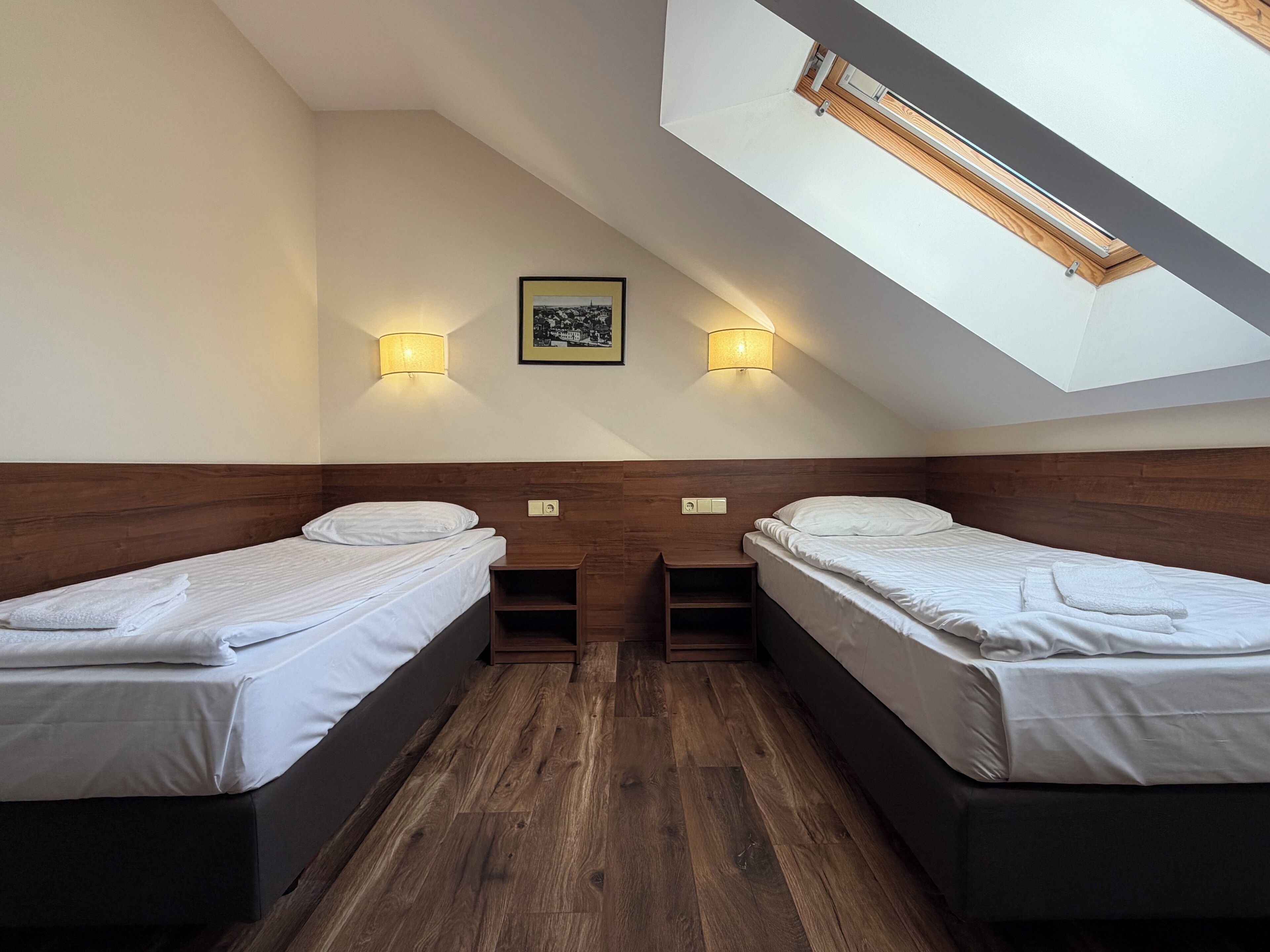 A room with two beds, wooden headboards, a skylight, and two wall-mounted lamps.