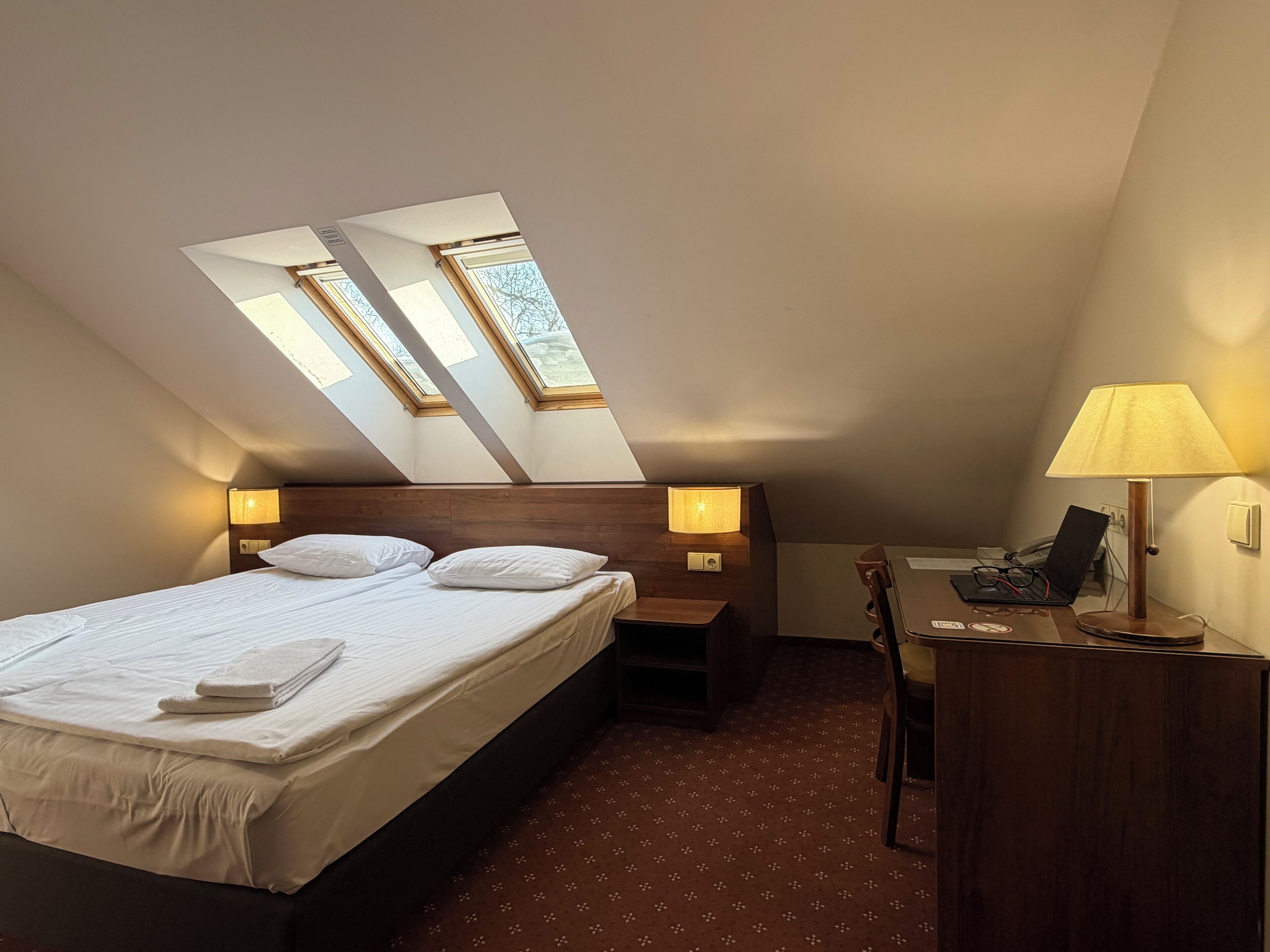 Ein Schlafzimmer mit einem Bett, zwei Nachttischlampen, einem Schreibtisch mit einem Laptop und einem Stuhl. Über dem Bett befinden sich Dachfenster.