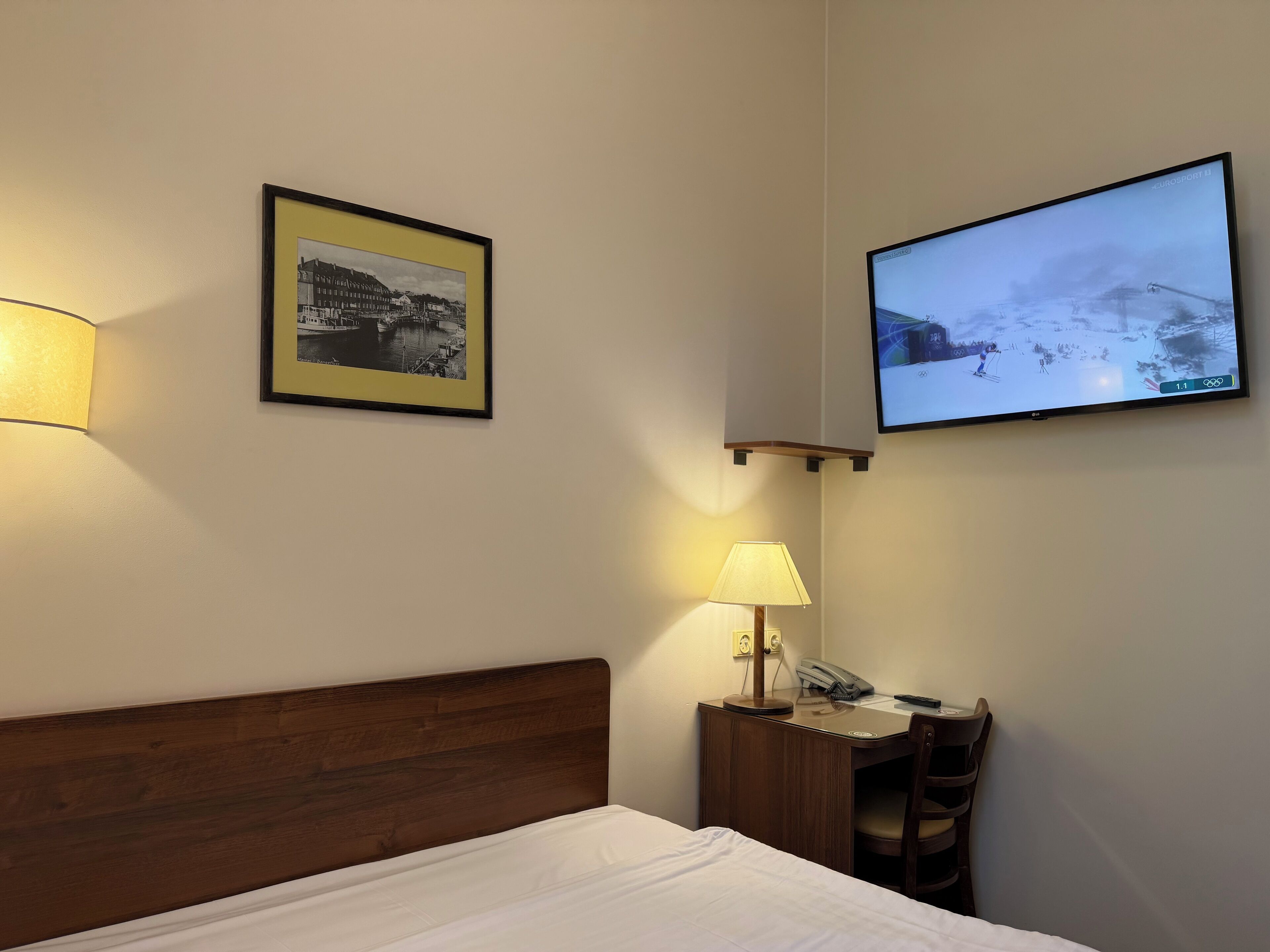 Une chambre d’hôtel avec un lit, un bureau doté d’une lampe, un tableau encadré au mur et un téléviseur diffusant une scène hivernale.