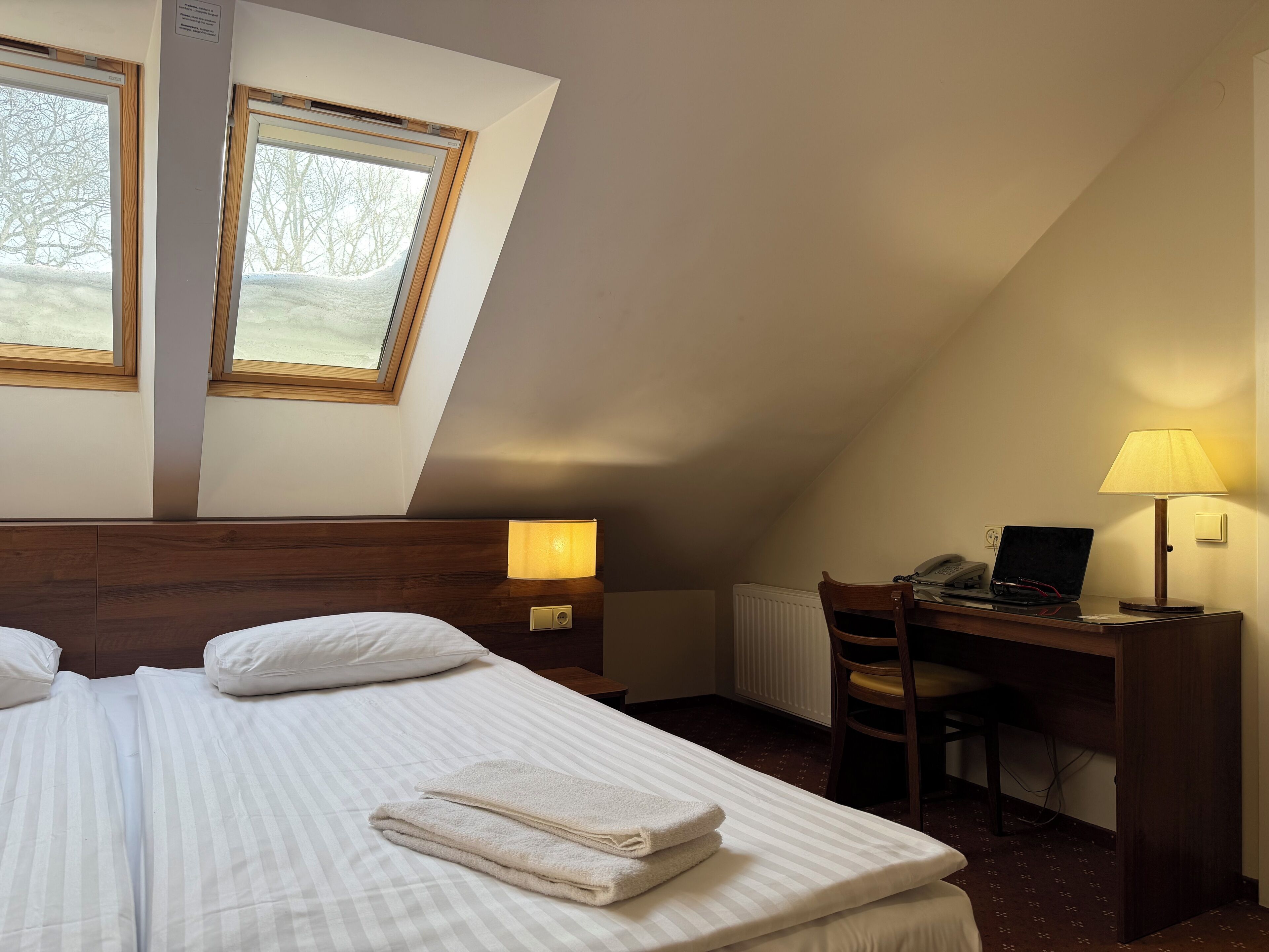 Habitación de hotel con cama, dos lámparas de noche, un escritorio con una computadora portátil, una silla y una ventana con vistas a árboles.