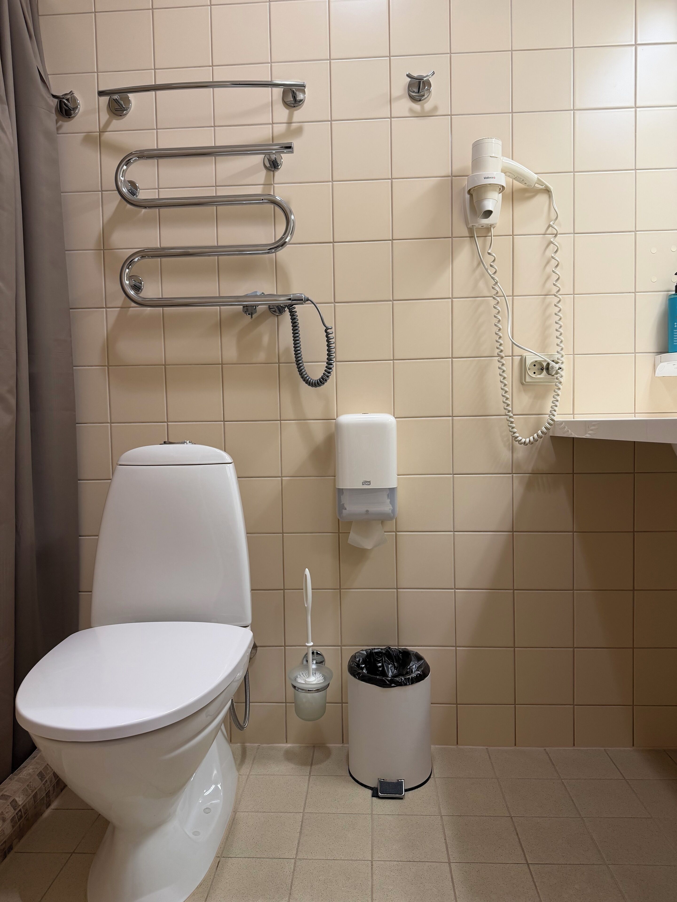 A bathroom with a toilet, grab bars, a hand dryer, a trash can, and a soap dispenser.