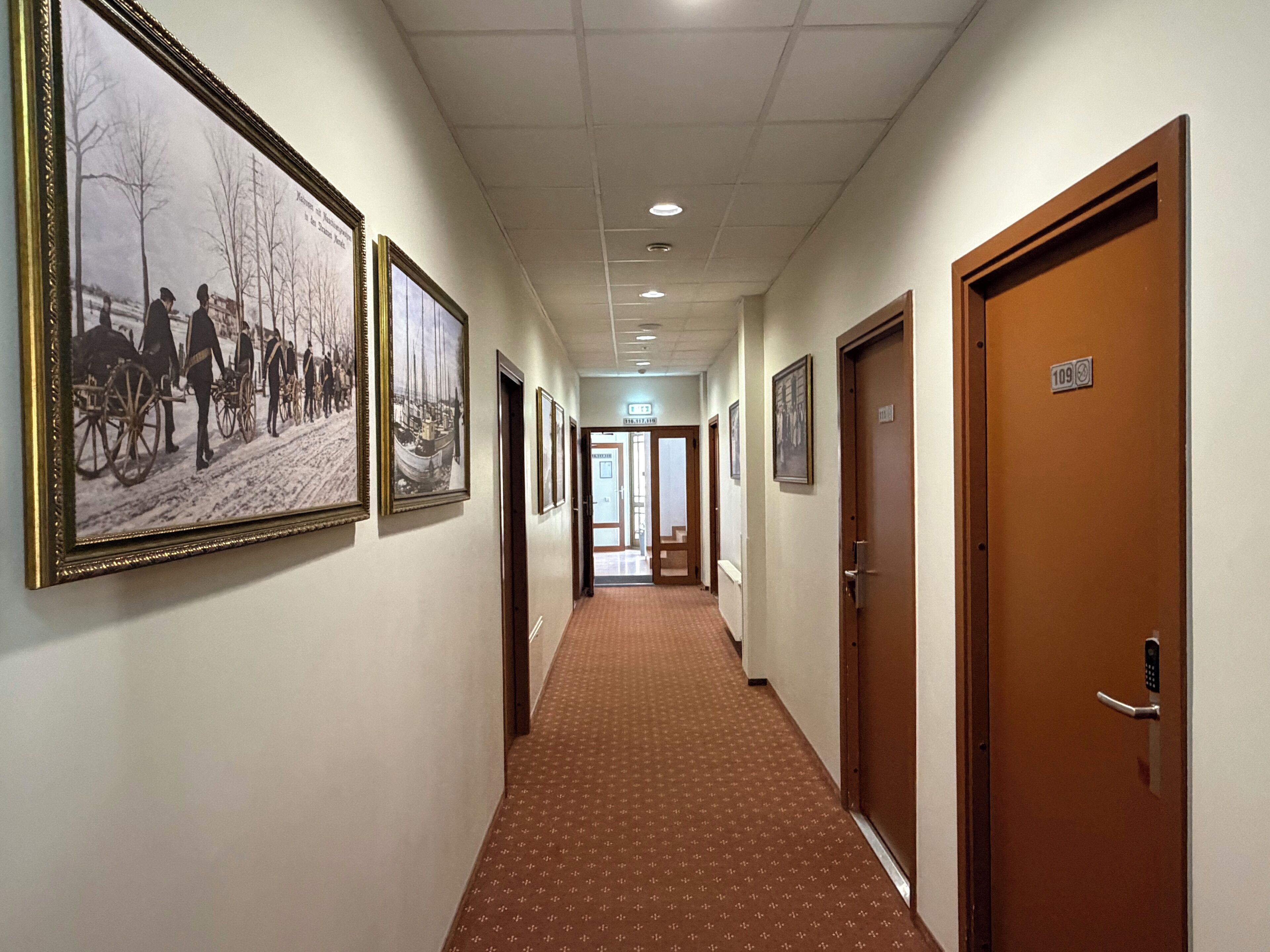 Un couloir d’hôtel orné de photographies historiques encadrées, avec un tapis à motifs et des portes numérotées 109 et 110.