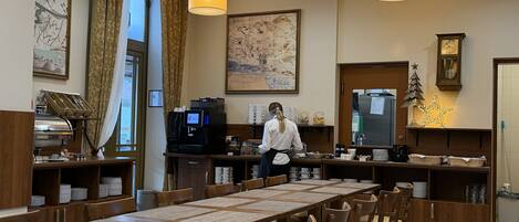 A hotel dining area with a long wooden table, chairs, a coffee machine, and a large map on the wall.