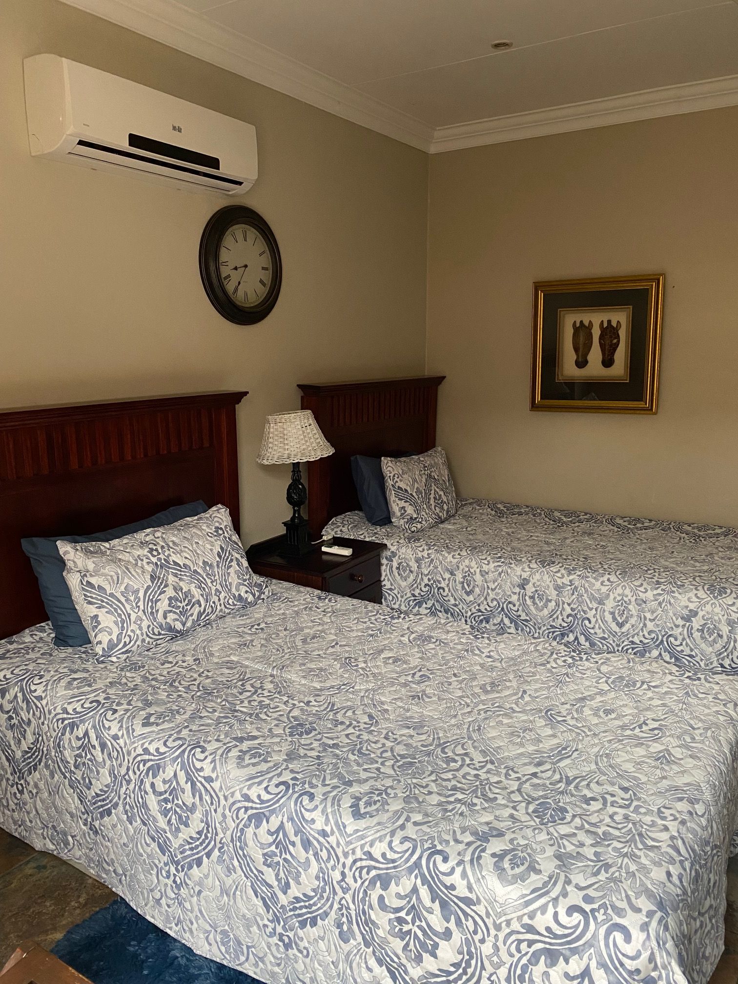 A hotel room with two beds, a clock, an air conditioner, a nightstand with a lamp, and a framed picture on the wall.