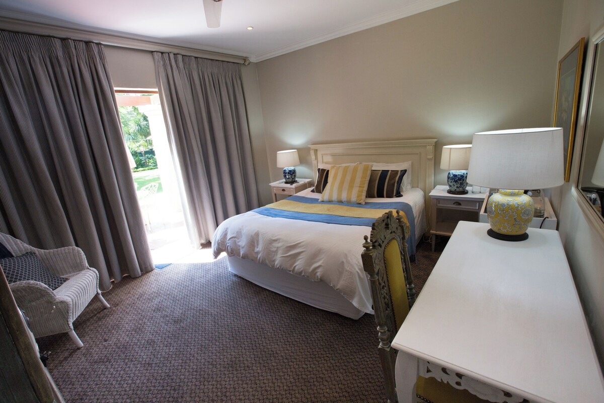 Habitación de hotel con cama, dos mesitas de noche con lámparas, una silla y un ventanal con cortinas.