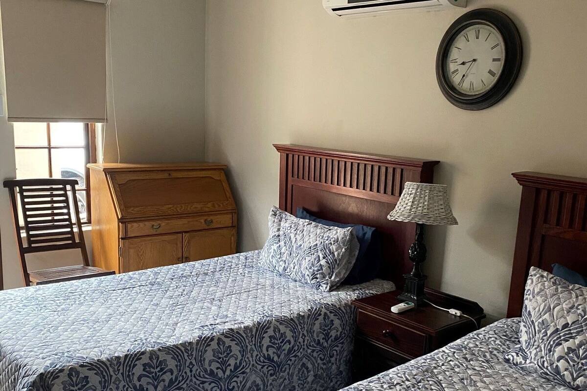 Hotellihuone, jossa on kaksi sänkyä, puinen lipasto, lamppu, kello ja ilmastointilaite.