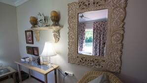 Een kamer met een grote, sierlijke spiegel, een rieten bank, een klein tafeltje met een lamp, en wandplanken met decoratieve objecten.