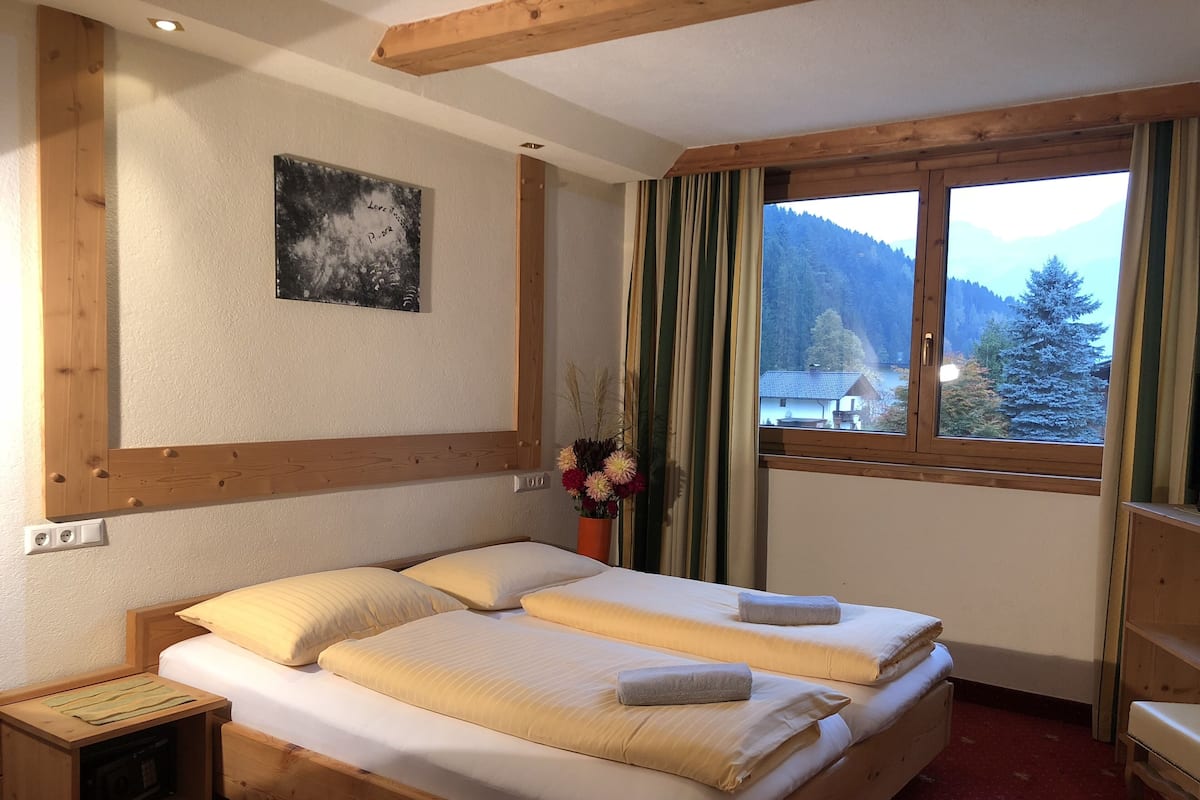 A double bed with two pillows and a vase of flowers, wooden headboard, a painting on the wall, and a window with a view of mountains.